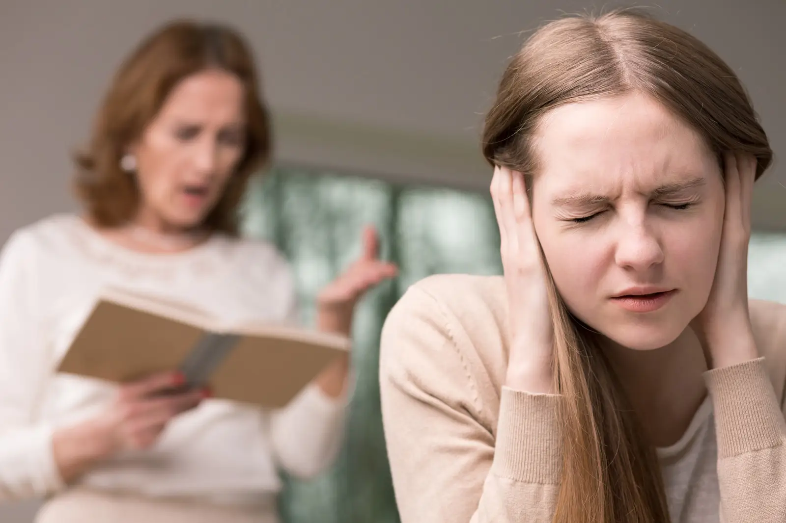 A photo depicting a young woman with a pained expression, holding her hands to her temples as if to block out sound, while an older woman in the background appears to be speaking to her in an agitated manner, possibly criticizing or arguing, which could suggest a stressful interaction related to Narcissistic Personality Disorder dynamics.