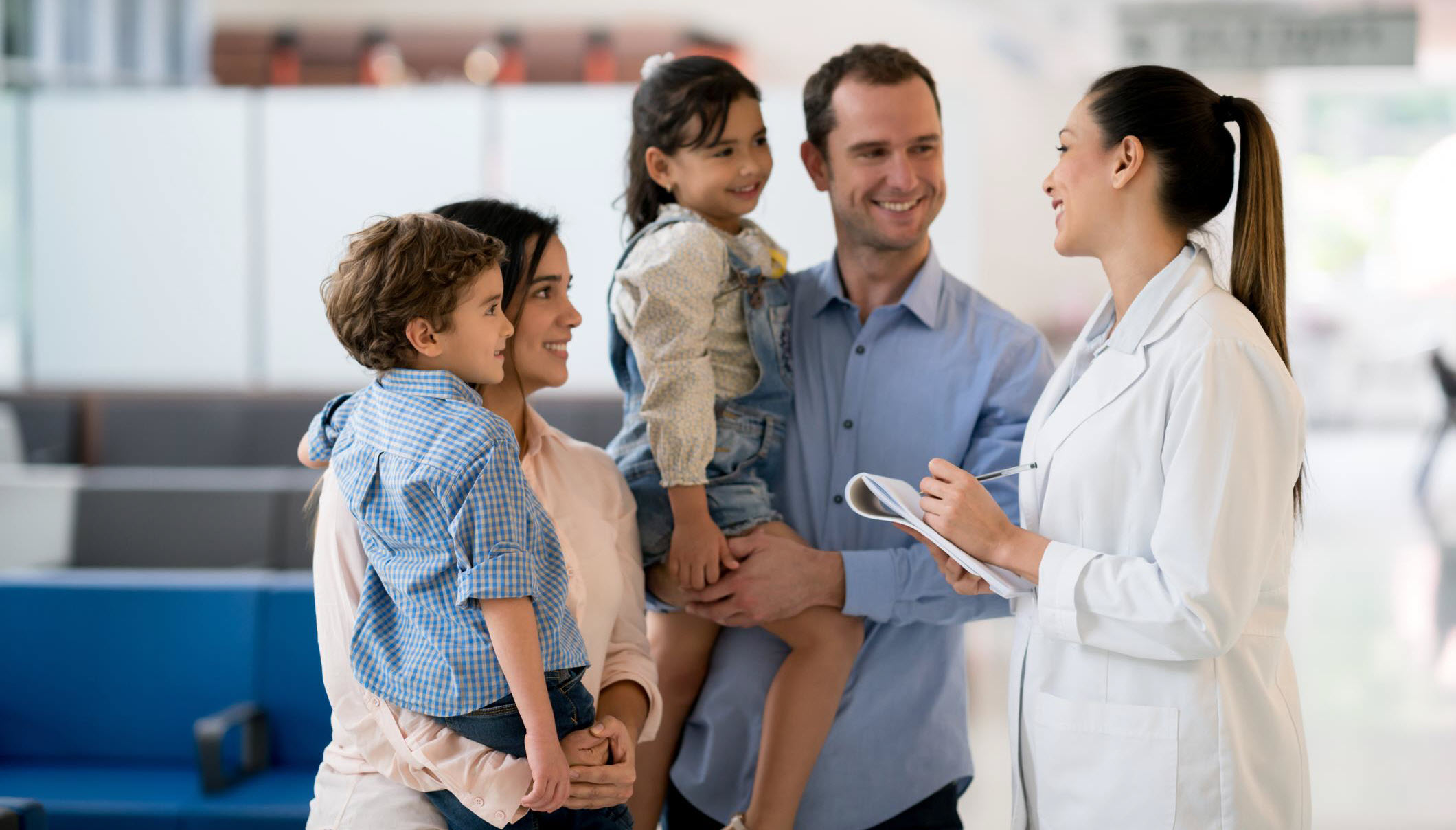A happy family consisting of a mother, father, and two children is engaging with a female healthcare professional, possibly during a family therapy session. The family is smiling and appears relaxed and comfortable in a clinical setting.